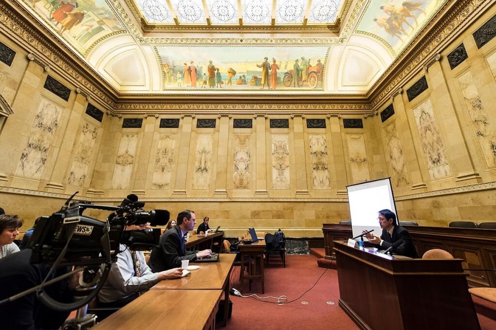 Legislators and staff gathered in an ornate hearing room at the state Capitol listen as UW virologist Yoshihiro Kawaoka explains his work to deter serious diseases such as Ebola.