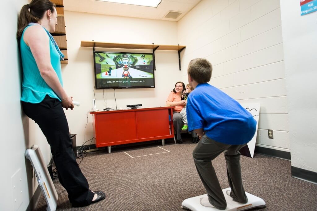 Xavier Hansen stands on a Wii Connect balance board and practices a Wii ski jump pose while mirroring his body position to match that of a video-game shape displayed on a computer monitor.