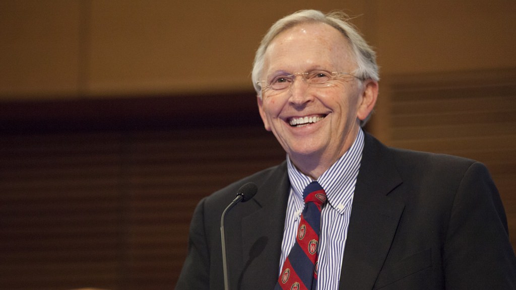 Grey-haired man in glasses, suit and red and blue tie, from behind podium, smiles towards his audience