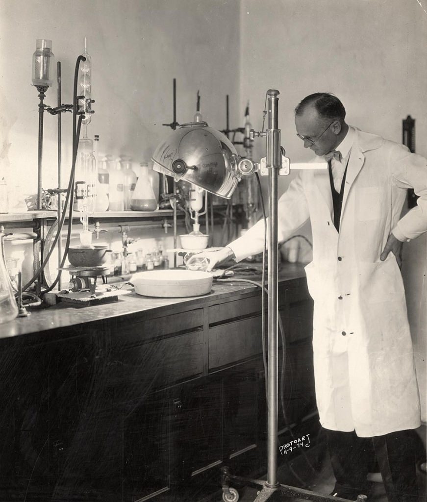 black and white photo of man in white lab coat, glasses, bow tie, pouring liquid into shallow container. Surrounding are various tools and containers, and a big floor lamp on wheels, consistant with old-fashioned lab