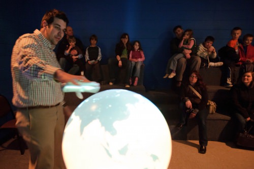 Referencing a 3-D weather globe displaying real-time and recorded satellite imagery, an outreach specialist with the Space Science and Engineering Center (SSEC), describes weather patterns on Earth to a group of families and visitors participating in Science Expeditions in 2011.