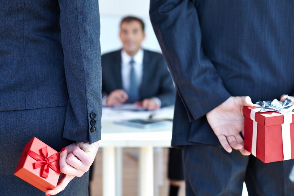 Photo: Two people in suits holding gift-wrapped boxes behind their backs, facing a man at a desk