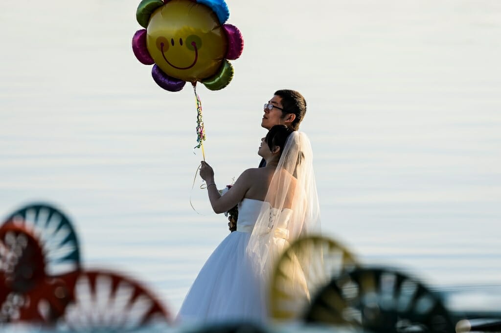 Photo: Couple in wedding clothes holding a smiley-face balloon by the lake