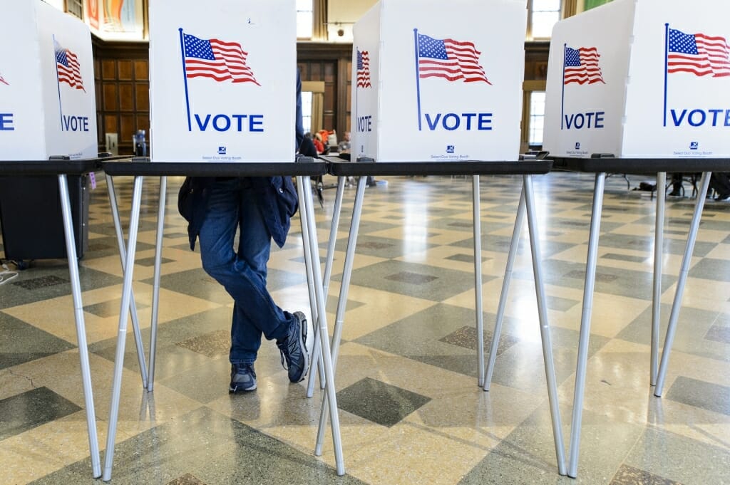 A person's leg's seen under a row of voting booths with American flags and the word 