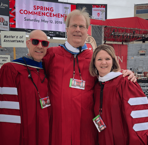 Photo: Argyle Wade in sunglasses, Kevin Helmkamp and Tonya Schmidt standing in red graduation gowns in front of Spring Commencement sign on jumbo-tron screen