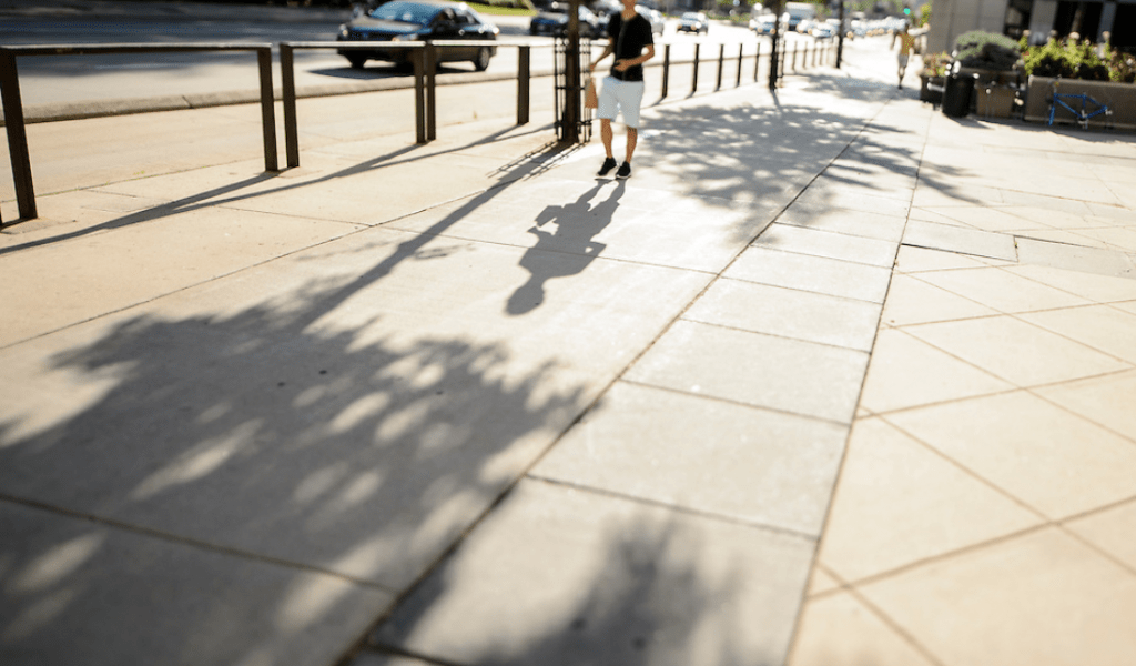 A passing pedestrian casts a shadow on a sidewalk along University Avenue near Grainger Hall of the University of Wisconsin–Madison campus as the sun rises during a summer morning.