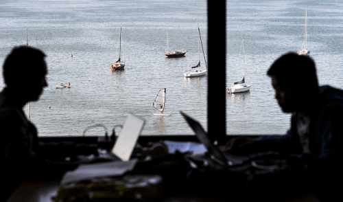 The juxtaposition of campus life during summer-session is seen as two silhouetted students study indoors next to a lakefront window in College Library at Helen C. White Hall.