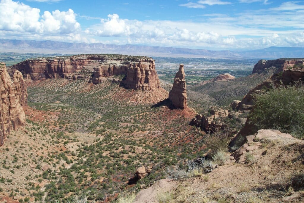 Colorado National Monument AltText: towers and mesas of brown rock in the midground with shrubby desert ground taking up most of the foreground