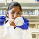 Photo: Molugu in blue gloves and white coat aiming pipette at lab dish