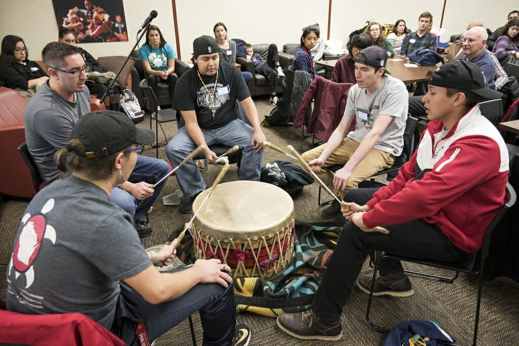 A number of students sit around a Native drum and bang it.