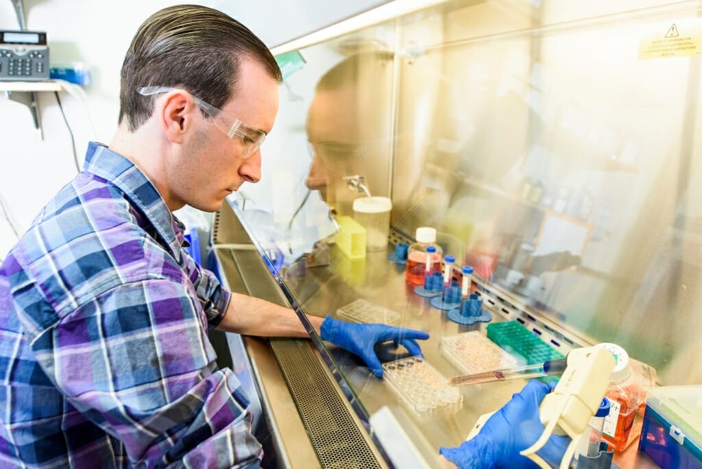 Photo: Simmons at lab bench in safety goggles and blue gloves