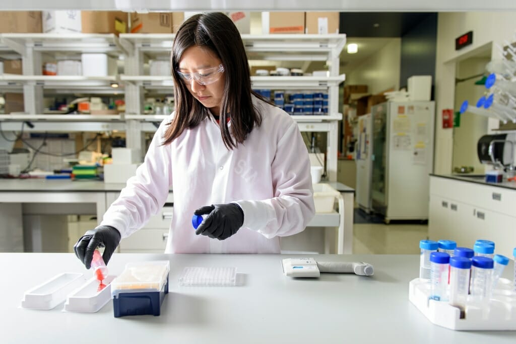 Photo: Xie in white lab coat and rubber gloves holding a sample at a lab bench