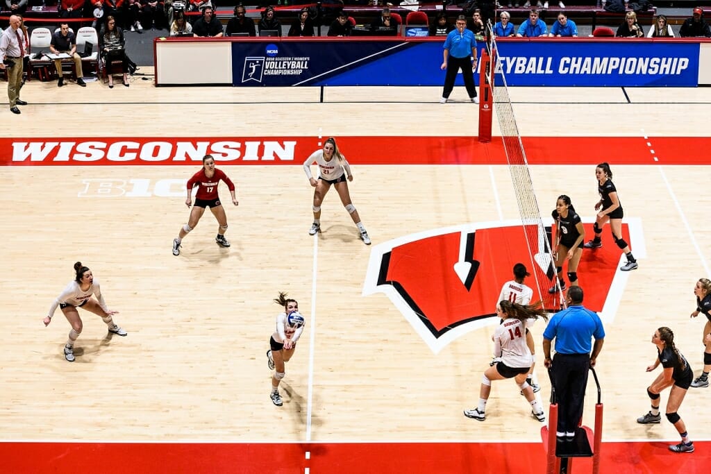 Volleyball setter Sydney Hilley, wearing no. 12, reaches to spike the ball as other players stand at the ready, in a full-court view.