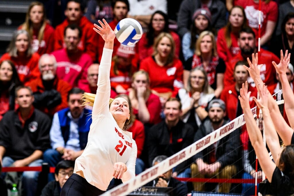 Player Grade Loberg, wearing No. 21, jumps high in the air to spike the ball over opposing players, as the crowd watches.