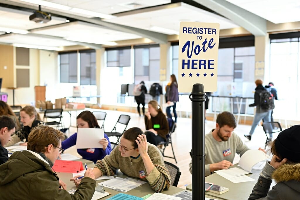 Photo: Students sitting at tables filling out forms, under a sign that says "Vote Here."