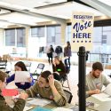 Photo: Students sitting at tables filling out forms, under a sign that says 