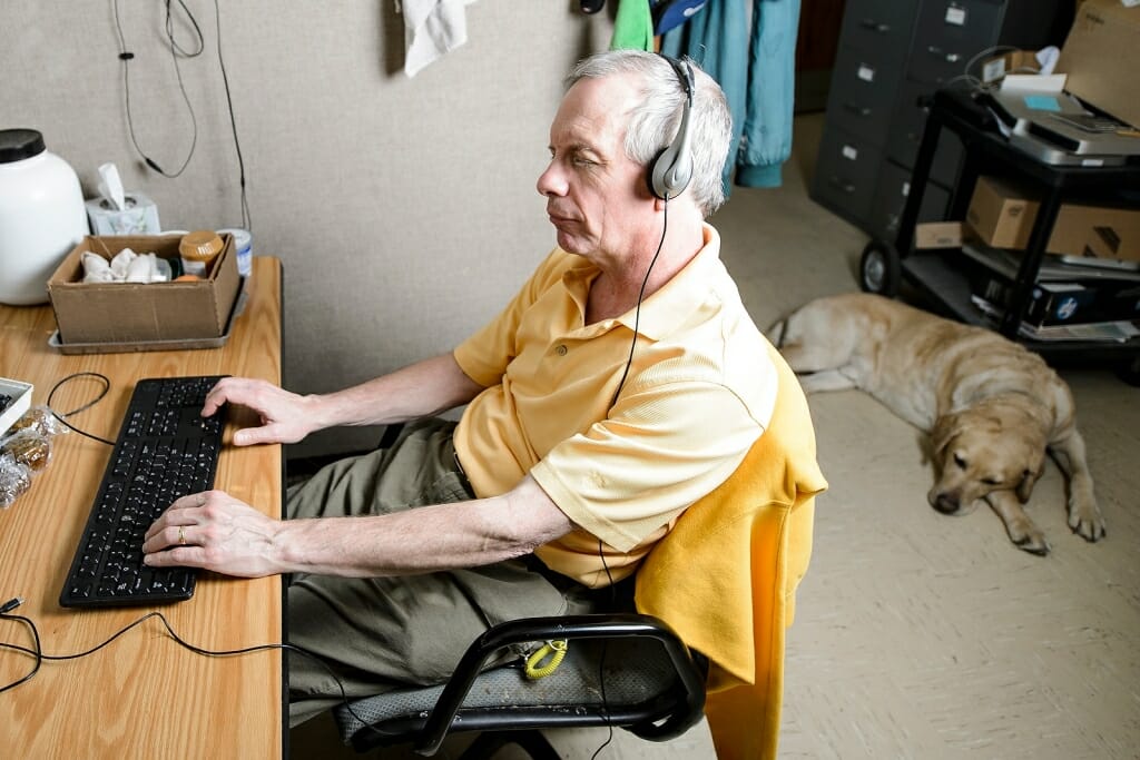 Photo: Man sitting at computer with headphones on and dog on floor behind