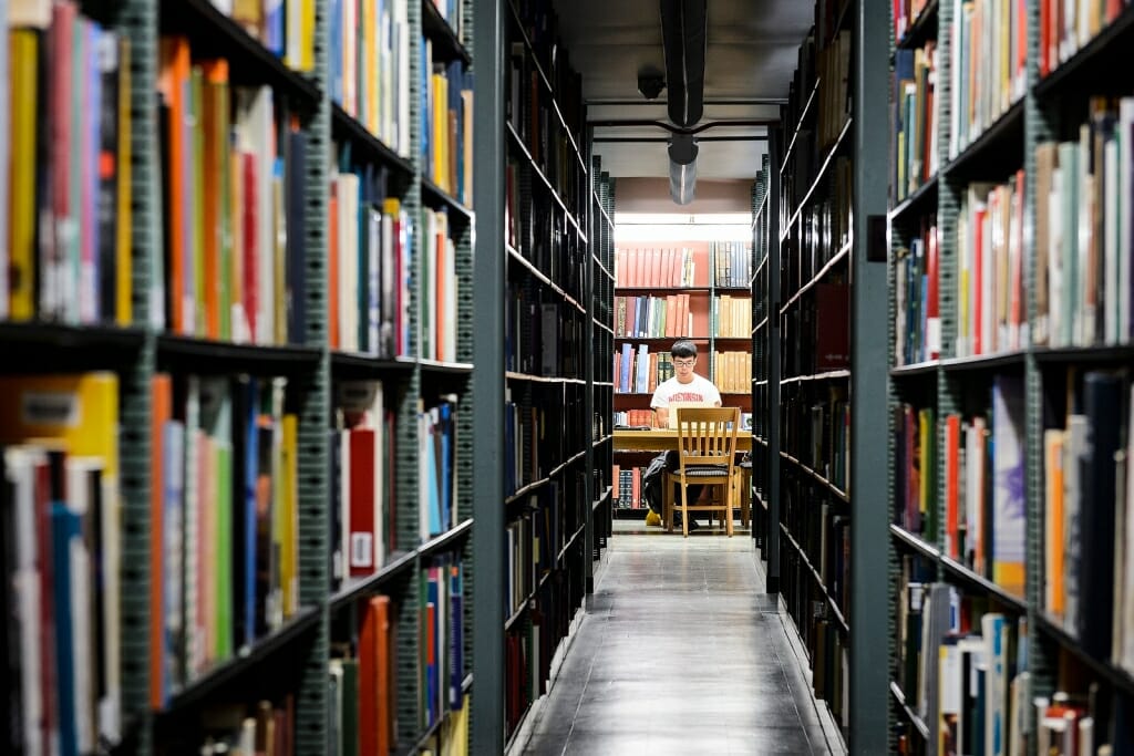 Photo: Person working on laptop as seen through two bookshelves