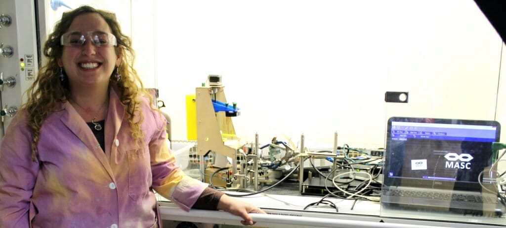 Photo: Woman with lab coat stands next to equipment.