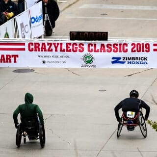 Photo: Two people in wheelchairs zoom down State Street below a banner that says Crazylegs Classic.