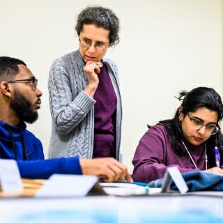 Photo: Susan standing and listening to student sitting at table