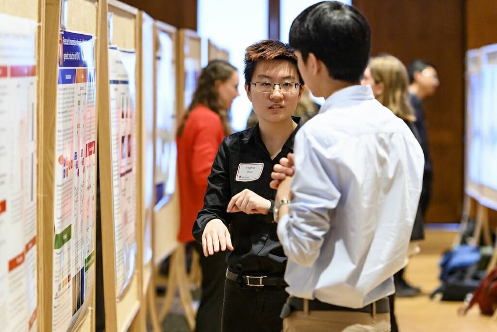 Photo: A student gestures and explains to an onlooker his project, next to a poster board.