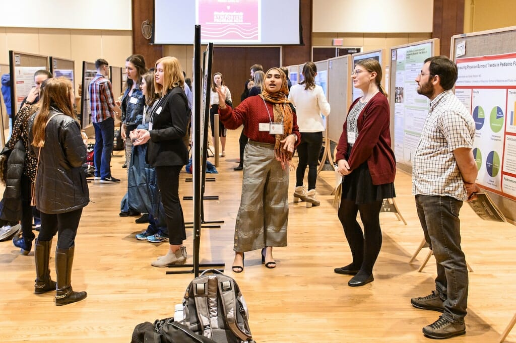 Photo: A series of student explain their projects in front of poster boards.