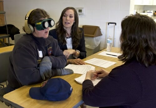 Photo: A man wearing goggles and earplugs talks with two women.