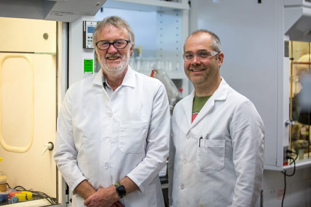 Photo; Two men in white lab coats smile in a lab.