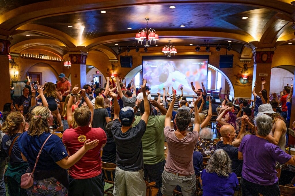 Photo of fans standing and cheering as they watch Lavelle being embraced by teammates on a large TV screen.