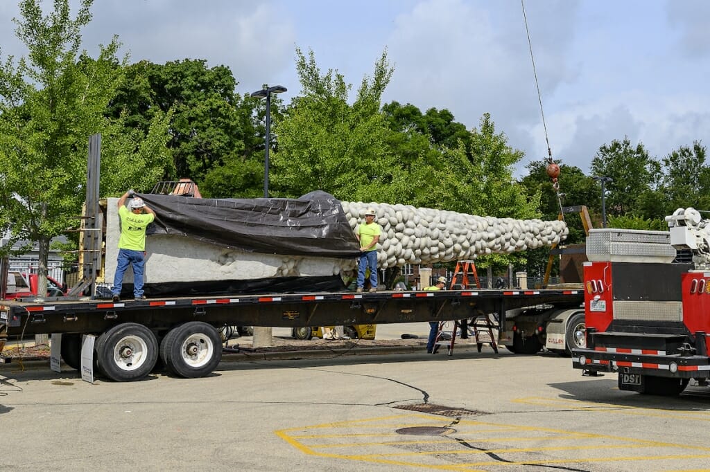 Photo: Workers pull a tarp over a sculpture laying down on a truck.