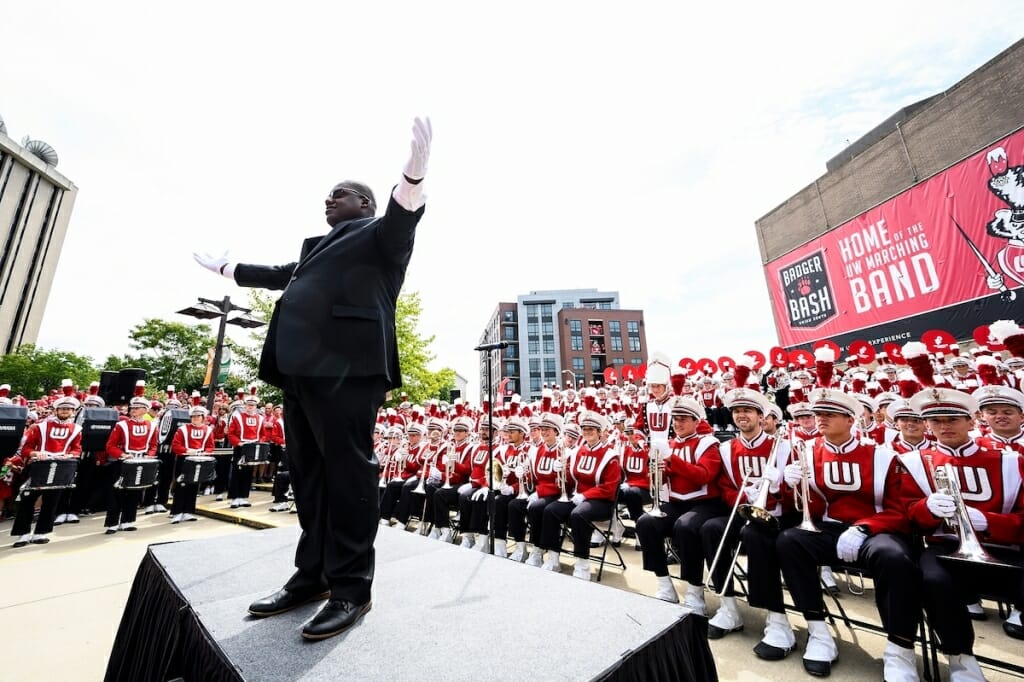 Photo: Pompey wearing dark suit and white gloves on platform with arms raised, surrounded by seated uniformed band members