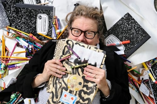 Photo of Lynda Barry holding an illustrated notebook, surrounded by notebooks and colored pens.