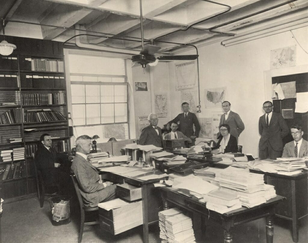 Photo: People sitting and standing in office around desks piled with papers