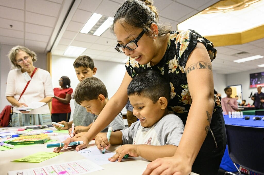 Photo: A boy draws on paper while his mother looks over his shoulder and smiles.