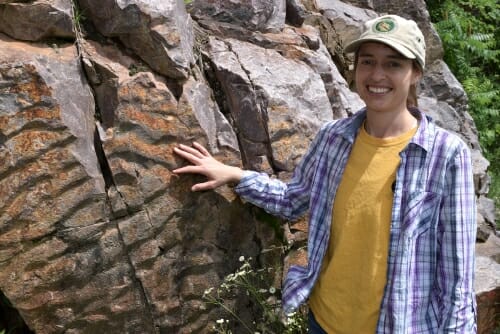 A woman stands in front of a large rock.