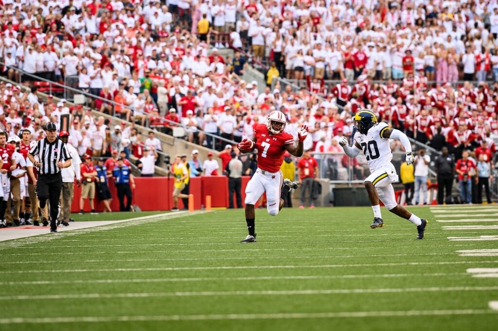 Photo: A Badger player runs, pursued by a Wolverine player, as fans cheer.