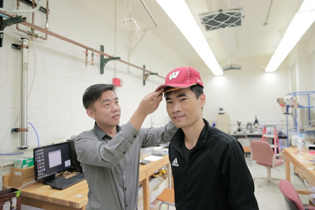 Photo: A man tries on a cap with a device in it, as another man helps.