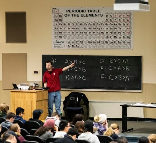 Photo: Bowman pointing at chalkboard below periodic table on wall