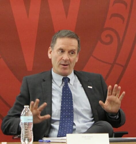 Photo: Green seated at table, speaking, in front of a large W crest on wall