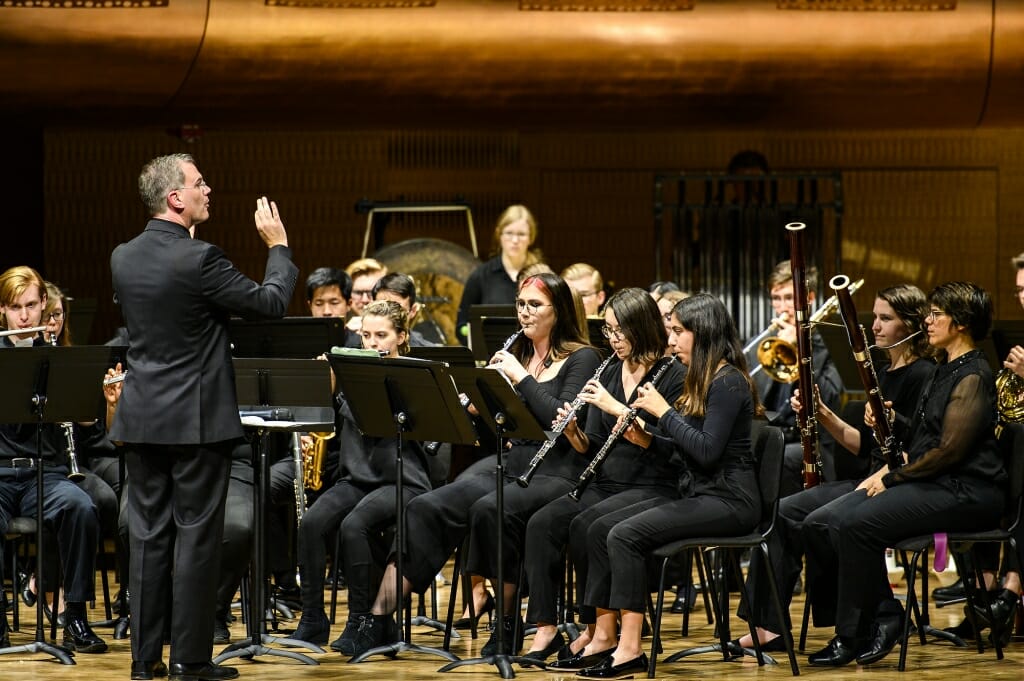 Photo: Conductor gesturing toward seated musicians playing wind instruments