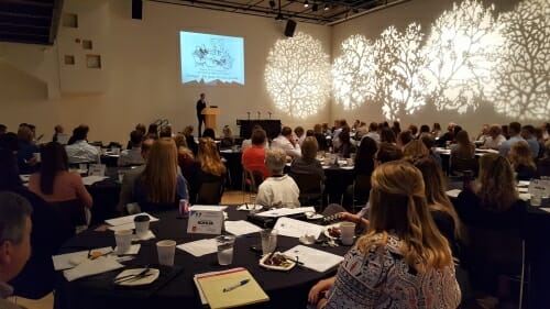 Photo: A woman addresses a room full of people.