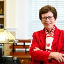 Portrait of Rebecca Blank wearing a W scarf standing in front of her desk with a lamp on it