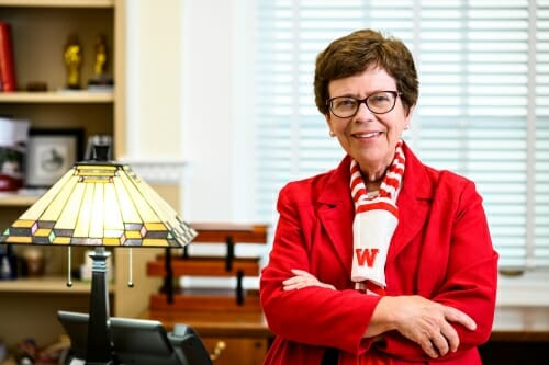 Portrait of Rebecca Blank wearing a W scarf standing in front of her desk with a lamp on it