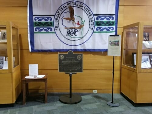 Photo: Plaque in front of Ho-Chunk Nation flag, flanked by display cases