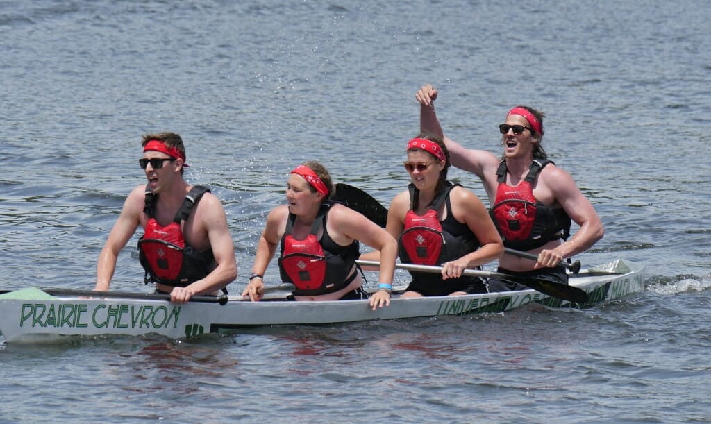 Photo: 4 people in life vests and red headbands rowing a concrete canoe named "Prairie Chevron" on a lake