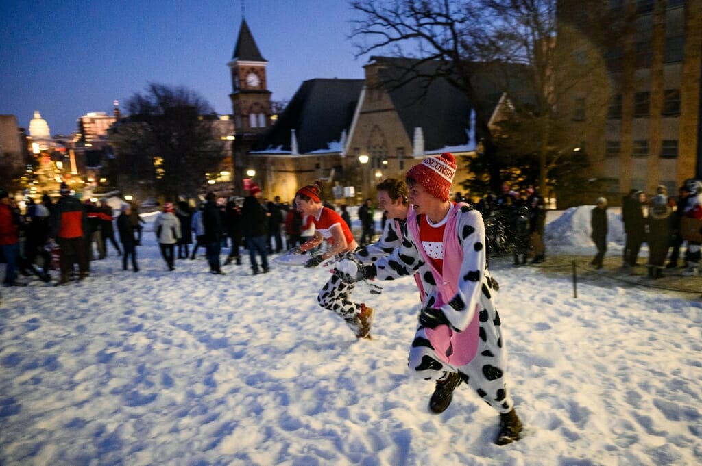 3 students in cow costumes running across the snow, with Capitol glowing in the distance