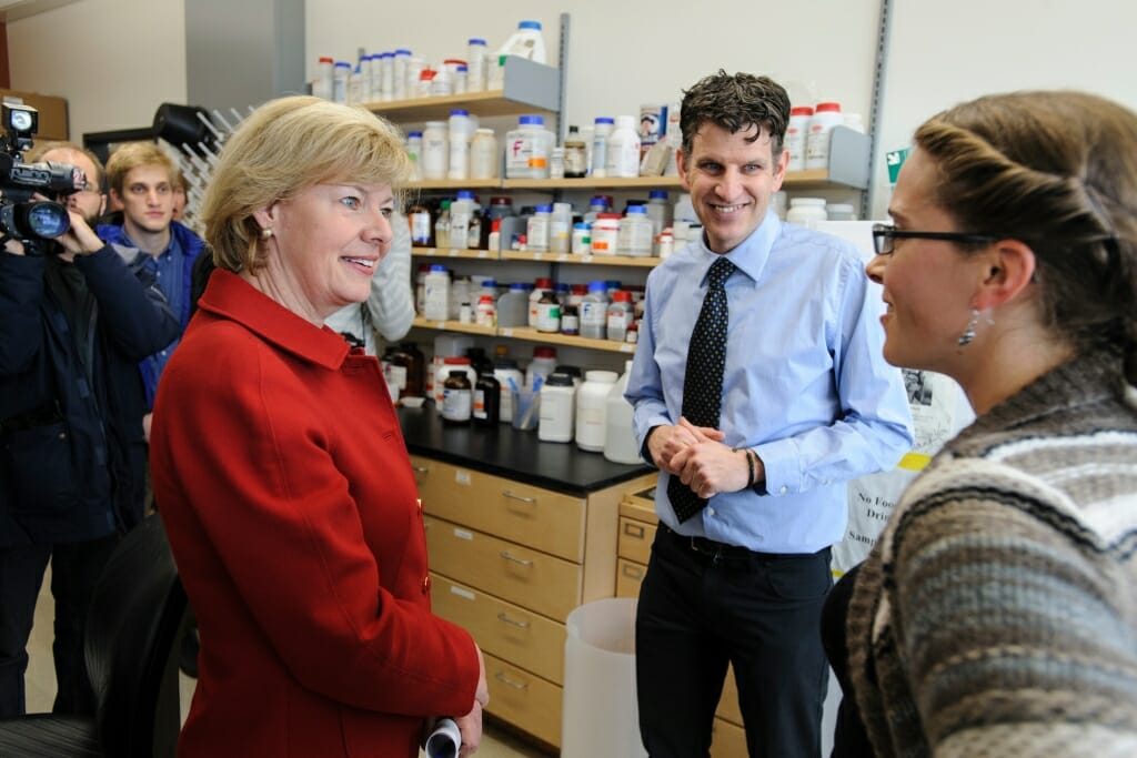 The senator and a man talk in a lab full of scientific equipment.