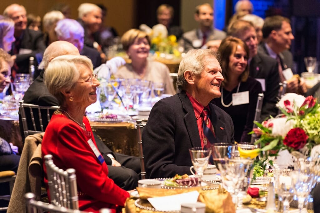 Tashia and John Morgridge sitting at a table smiling among other tables surrounded by people applauding