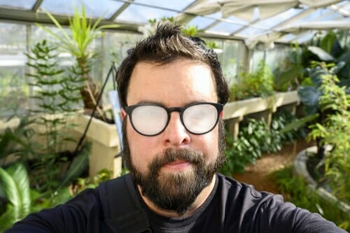 Bryce inside the greenhouse in front of green plants with his eyeglasses fogged over